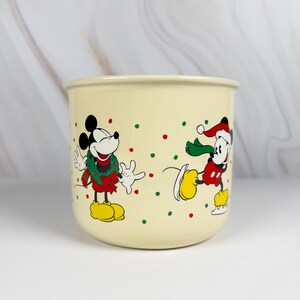 May include: Cream-colored ceramic mug featuring Mickey Mouse in festive holiday attire. One Mickey is wearing a wreath, the other is ice skating. Red and green polka dots decorate the mug, adding to the holiday theme.