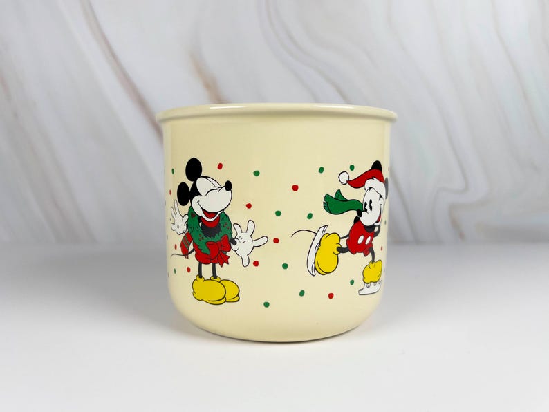 May include: Cream-colored ceramic mug featuring Mickey Mouse in festive holiday attire. One Mickey is wearing a wreath, the other is ice skating. Red and green polka dots decorate the mug, adding to the holiday theme.