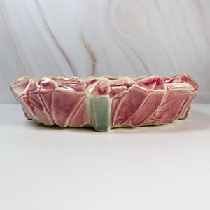 May include: A pink ceramic planter with a leaf-like design. The planter has a rectangular shape with a scalloped rim and a light green accent on the side. The planter is likely for indoor use.