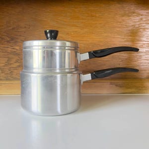 May include: A vintage aluminum double boiler with a black handle and lid knob. The pot has a shiny, silver finish and is designed for cooking delicate foods. The handles are curved for easy gripping.