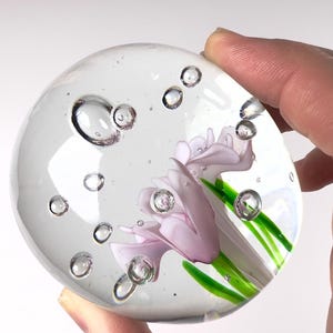 May include: A clear glass paperweight with a delicate floral design. The paperweight features a light purple flower with green stems and leaves, encased in a transparent sphere with air bubbles. The paperweight is held by a hand.