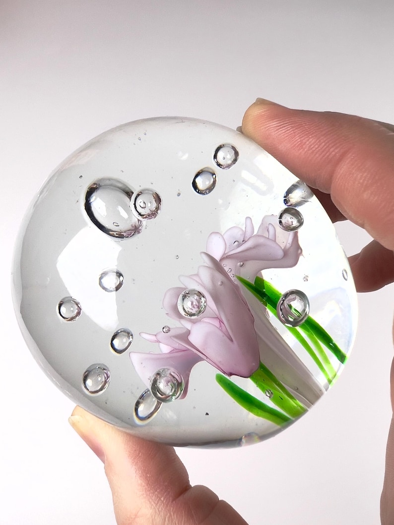 May include: A clear glass paperweight with a delicate floral design. The paperweight features a light purple flower with green stems and leaves, encased in a transparent sphere with air bubbles. The paperweight is held by a hand.