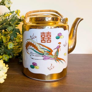 May include: A gold teapot with a bamboo handle and a white porcelain body. The teapot is decorated with a phoenix design and the Chinese character for "double happiness" in red.