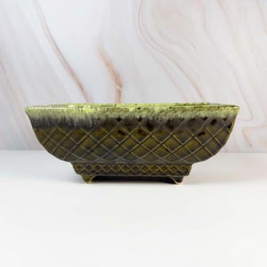 May include: A rectangular, olive-green ceramic planter with a diamond pattern and small feet. The top edge has a lighter green and brown glaze. The planter is suitable for indoor or outdoor use and is a decorative item.