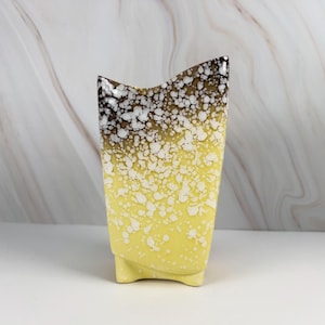 May include: A tall, ceramic vase with a unique shape and speckled design. The vase transitions from a dark brown top to a yellow base, with white speckles throughout. It has a flared top and a rectangular base with small feet.