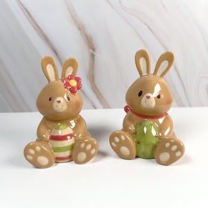 May include: Two ceramic Easter bunny figurines. One bunny has a red flower and holds a striped egg. The other bunny has a red bow and holds a green egg. Both bunnies are tan with white and brown accents.