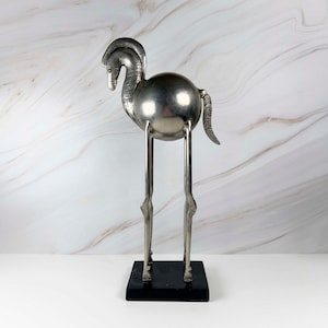May include: A silver-toned metal sculpture of a stylized, long-legged creature with a spherical body, a curved neck, and a textured tail. The figure stands on a rectangular black base, featuring intricate etched patterns along the neck area.