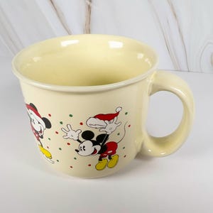 May include: Cream-colored ceramic mug featuring Mickey Mouse and Minnie Mouse in Santa hats, with red, green, and yellow accents. The mug has a large handle and is decorated with festive polka dots, perfect for holiday beverages.