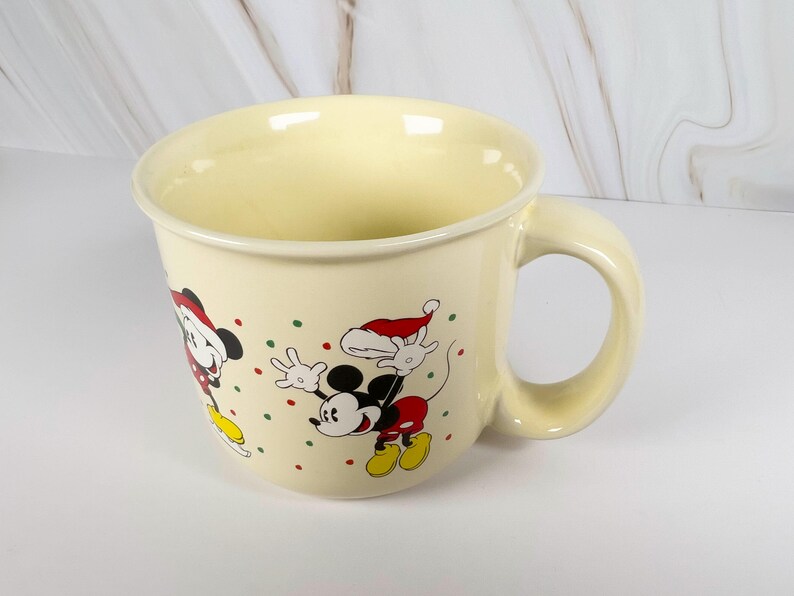 May include: Cream-colored ceramic mug featuring Mickey Mouse and Minnie Mouse in Santa hats, with red, green, and yellow accents. The mug has a large handle and is decorated with festive polka dots, perfect for holiday beverages.