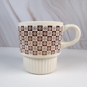 May include: A cream-colored ceramic mug featuring a brown checkered pattern with small floral motifs in each square. The upper portion of the mug is cylindrical, while the lower section has vertical ribbed detailing and a curved handle.
