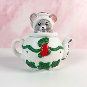May include: A white ceramic teapot with a Christmas theme. The teapot features green holly and red berry accents, a red stocking, and a gray mouse figurine on the lid. The teapot has a handle and a spout.