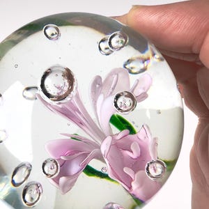 May include: A clear glass paperweight with a delicate pink flower encased inside. The paperweight has several air bubbles suspended within the glass. The flower has green leaves and is surrounded by the clear glass.
