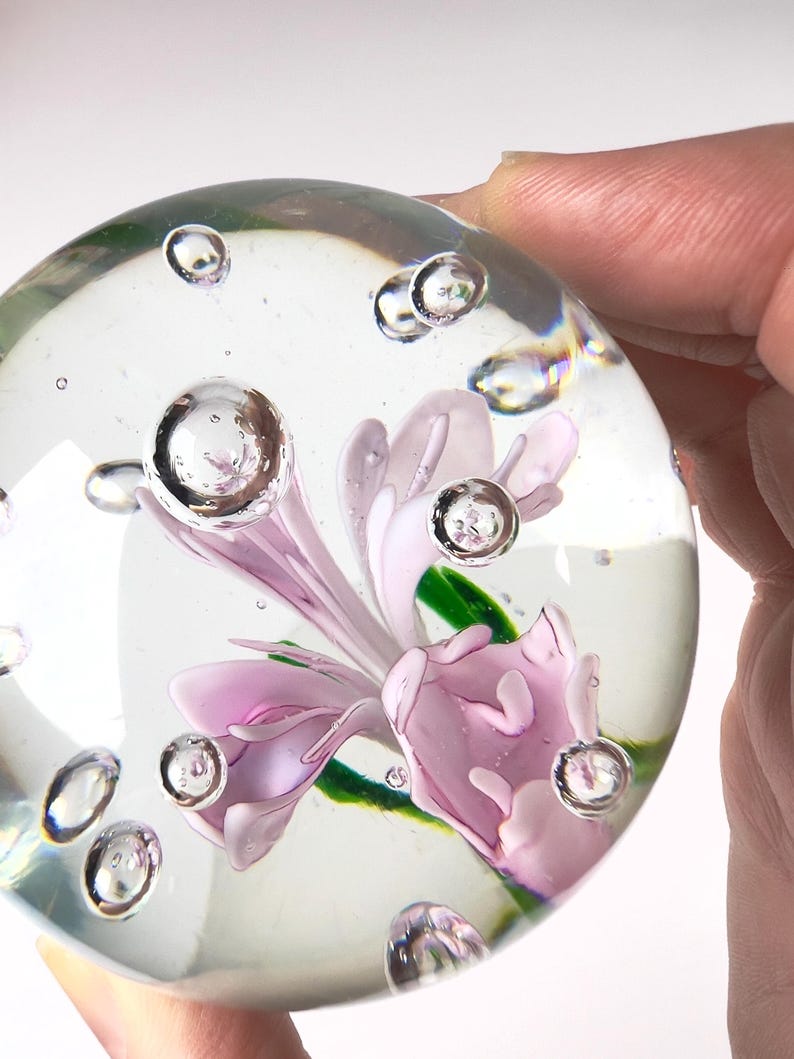 May include: A clear glass paperweight with a delicate pink flower encased inside. The paperweight has several air bubbles suspended within the glass. The flower has green leaves and is surrounded by the clear glass.