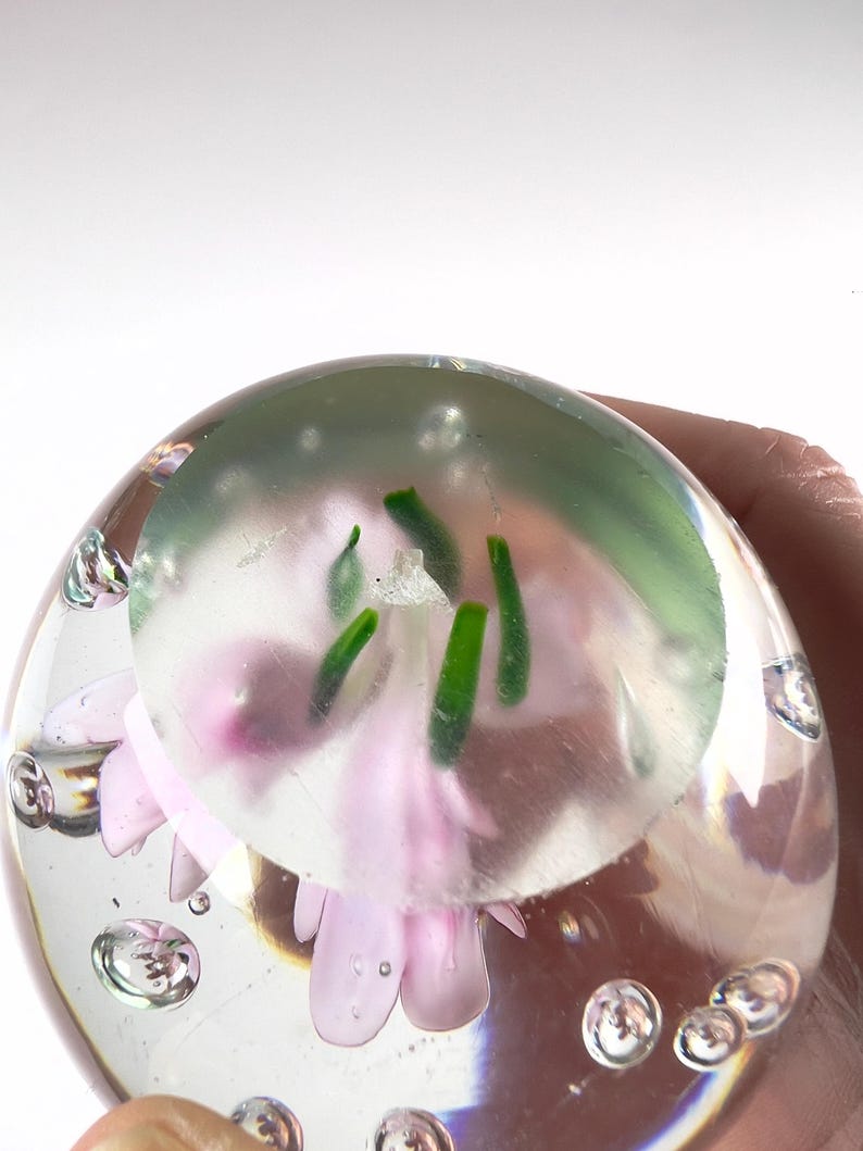May include: A clear glass paperweight with a pink flower and green stems encased inside. The paperweight is round with air bubbles throughout. The flower has delicate petals and is centered within the glass sphere.