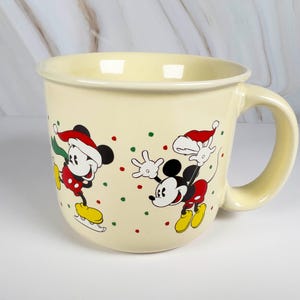May include: Cream-colored ceramic mug featuring Mickey Mouse in a Santa hat, ice skating. The mug has a large handle and is decorated with red, green, and yellow accents. A festive holiday-themed mug.