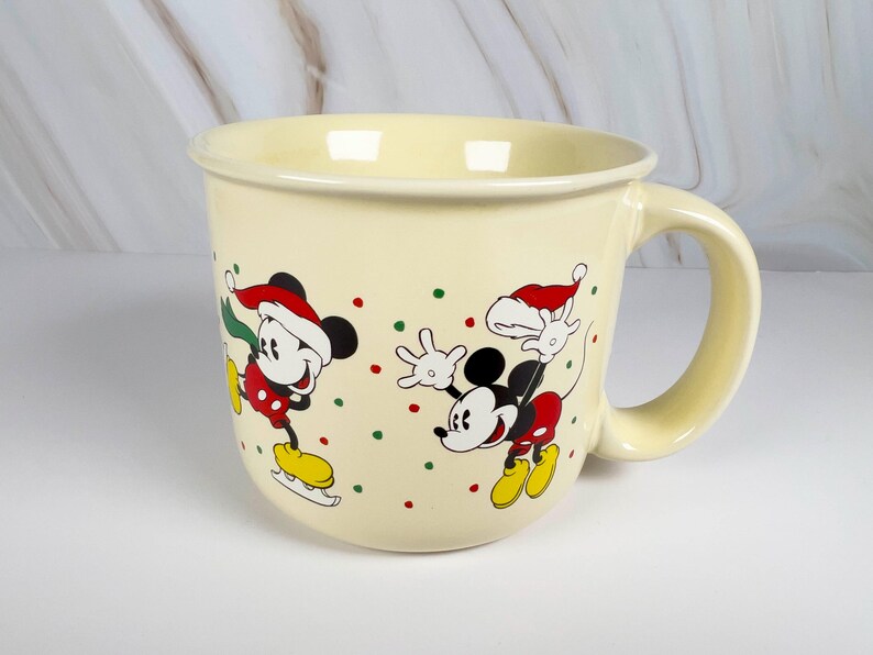 May include: Cream-colored ceramic mug featuring Mickey Mouse in a Santa hat, ice skating. The mug has a large handle and is decorated with red, green, and yellow accents. A festive holiday-themed mug.