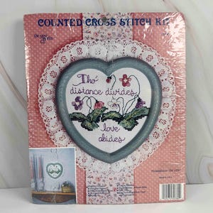 May include: A counted cross stitch kit featuring a heart-shaped design with the text "Tho' distance divides, love abides." The design includes flowers and greenery. The kit is packaged in a pink and white polka dot background with a lace doily.