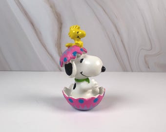 May include: A white Snoopy figurine with black ears and a green collar emerging from a cracked pink and purple Easter egg with blue polka dots, while a small yellow Woodstock bird figure sits perched on top of the egg shell.