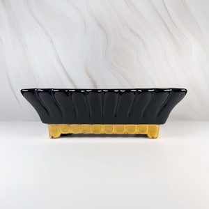 May include: A rectangular ceramic planter featuring a glossy black finish with a scalloped, fluted upper edge. The base of the vessel is finished in a metallic gold tone with a repeating vertical ribbed pattern, supported by four small feet.