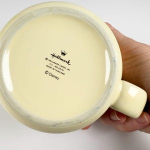 May include: Cream-colored ceramic mug with a rounded base and a handle. The bottom of the mug features the Hallmark logo, the text "Hallmark Cards Inc. K.C., MO 64141 Made in Thailand" and the Disney logo.
