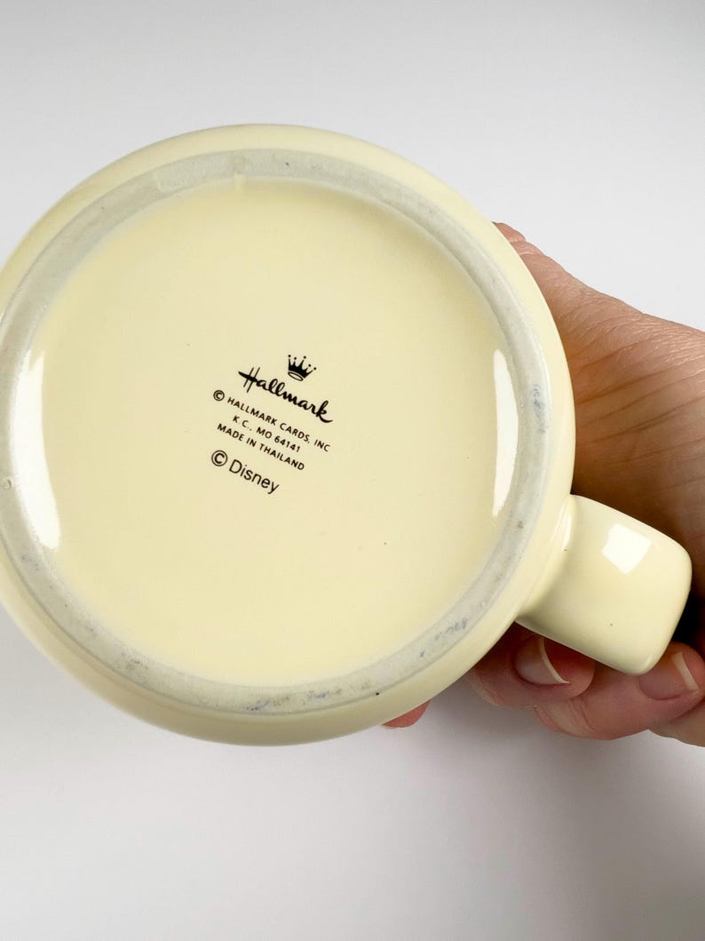 May include: Cream-colored ceramic mug with a rounded base and a handle. The bottom of the mug features the Hallmark logo, the text "Hallmark Cards Inc. K.C., MO 64141 Made in Thailand" and the Disney logo.