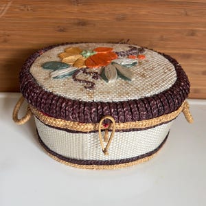 May include: A round, woven basket with a lid. The lid is decorated with an embroidered floral design in shades of orange, yellow, and green. The basket has a dark brown braided trim and handles. The basket is secured with a wooden clasp.