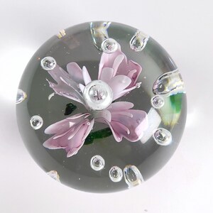 May include: A clear glass paperweight with a delicate, light purple flower encased within. The flower has multiple petals and a silver center. Bubbles are suspended in the glass, adding to the decorative appeal.