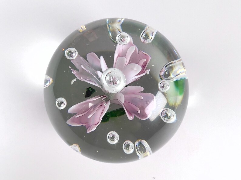 May include: A clear glass paperweight with a delicate, light purple flower encased within. The flower has multiple petals and a silver center. Bubbles are suspended in the glass, adding to the decorative appeal.
