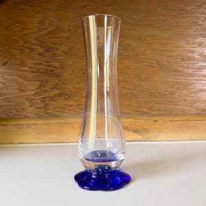 May include: A clear glass vase with a flared shape and a vibrant blue base. The vase has a slender, elegant design, perfect for displaying flowers or as a decorative piece. The blue base adds a pop of color and visual interest to the overall aesthetic.