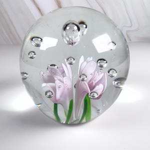 May include: A clear glass paperweight with a floral design. The paperweight features two light purple flowers with green stems encased in a clear glass sphere. Air bubbles are suspended throughout the glass.