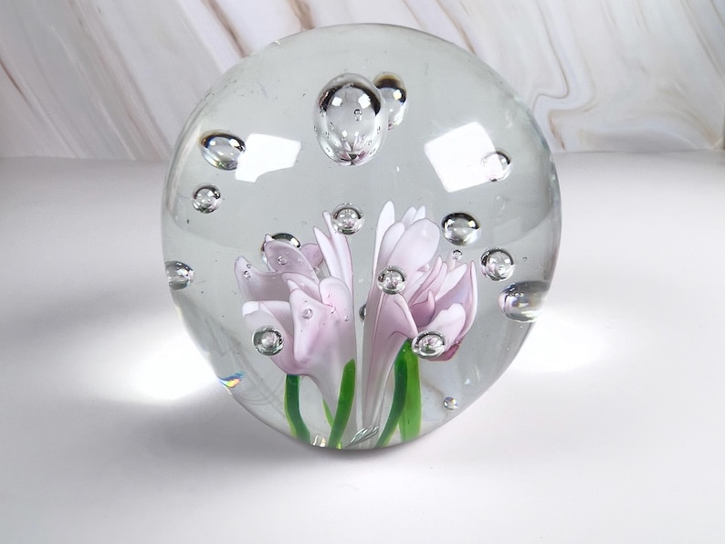May include: A clear glass paperweight with a floral design. The paperweight features two light purple flowers with green stems encased in a clear glass sphere. Air bubbles are suspended throughout the glass.
