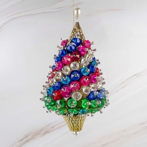 May include: A cone-shaped hanging ornament covered in multi-colored faceted beads and clear crystals. The design features rows of blue, pink, red, and green beads with a gold-toned textured base and a small white pearl-like bead at the top.