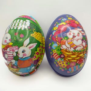May include: Two vintage Easter egg-shaped tins with colorful illustrations of bunnies and flowers. One tin is green with a bunny holding a list that says "Easter List" and the other tin is purple with a bunny holding an Easter egg in a basket.