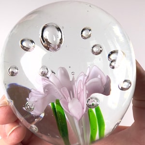 May include: A clear glass paperweight with a light purple flower and green stems encased inside. The paperweight is round with air bubbles throughout. The paperweight is held in a hand.