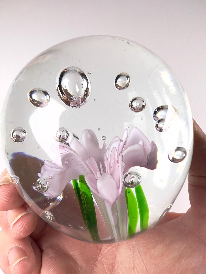 May include: A clear glass paperweight with a light purple flower and green stems encased inside. The paperweight is round with air bubbles throughout. The paperweight is held in a hand.