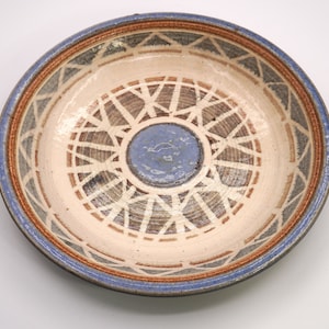Puede incluir: Un plato de cerámica con un diseño geométrico azul, marrón y blanco. El plato tiene un borde elevado y un círculo central con un esmalte azul.