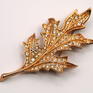 May include: A gold-toned leaf brooch with clear rhinestones. The brooch is shaped like a leaf with a pointed tip and a stem.