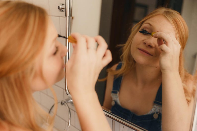 May include: A person applying mascara in front of a mirror. The person is wearing denim overalls. The bathroom has white tile and a chrome towel rack. The mascara wand is yellow and black.