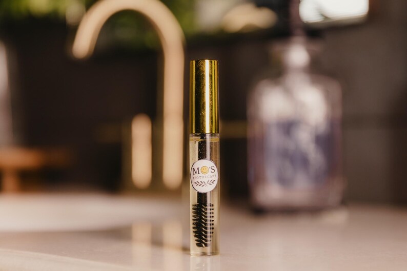 May include: A clear glass tube with a gold cap and a black mascara brush. The tube has a white label with the text "M O'S APOTHECARY" and a circular design. The background is blurred, with a gold faucet and a bottle.