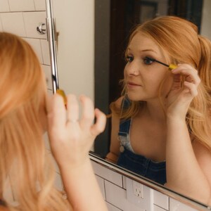 May include: A person applying mascara in front of a mirror. The person has long, reddish-blonde hair and is wearing a denim overall. The mirror is in a bathroom with white tile.