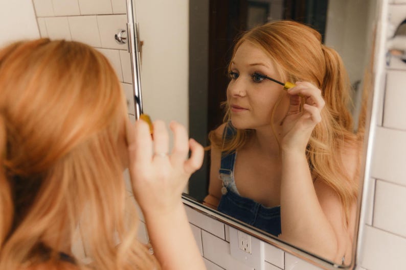 May include: A person applying mascara in front of a mirror. The person has long, reddish-blonde hair and is wearing a denim overall. The mirror is in a bathroom with white tile.
