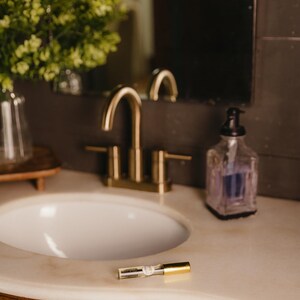 May include: A bathroom vanity with a gold faucet, white sink, and a clear soap dispenser. A mascara tube with gold accents rests on the marble countertop. The vanity has ornate wooden cabinets.