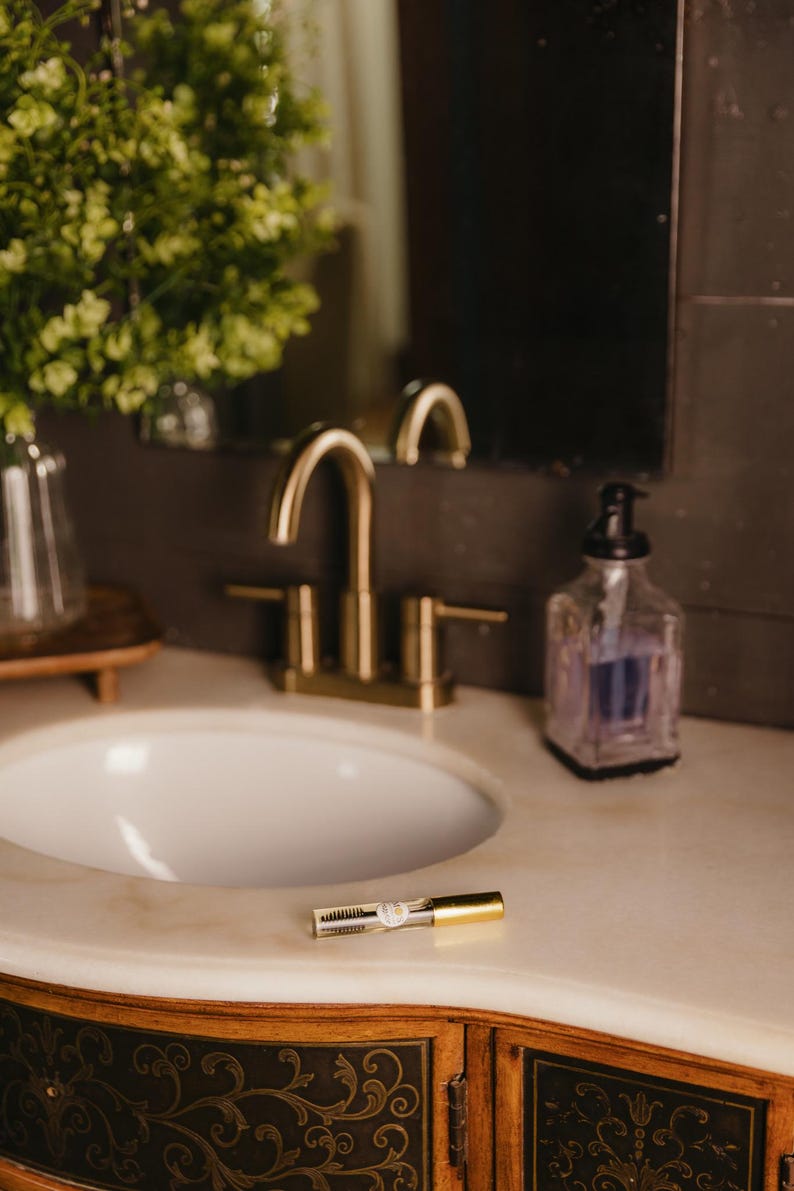 May include: A bathroom vanity with a gold faucet, white sink, and a clear soap dispenser. A mascara tube with gold accents rests on the marble countertop. The vanity has ornate wooden cabinets.