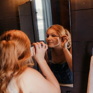 May include: A person applying mascara in front of a mirror in a bathroom. The mirror is framed by dark wood paneling. The person is wearing denim overalls. A light fixture with three bulbs is above the mirror.