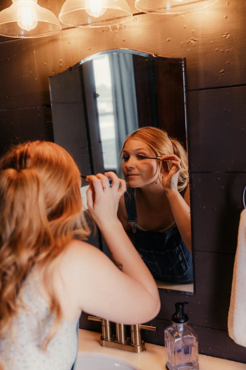 May include: A person applying mascara in front of a mirror in a bathroom. The mirror is framed by dark wood paneling. The person is wearing denim overalls. A light fixture with three bulbs is above the mirror.