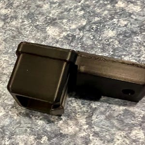 May include: A black, rectangular trailer hitch receiver with a square opening and a connecting piece. The receiver has a hole for a pin and is made of a durable material.