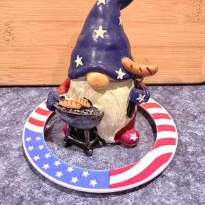 May include: A patriotic gnome figurine with a blue hat adorned with white stars, holding a miniature grill with hot dogs and a sausage. The base is a ring with red and white stripes and blue stars.