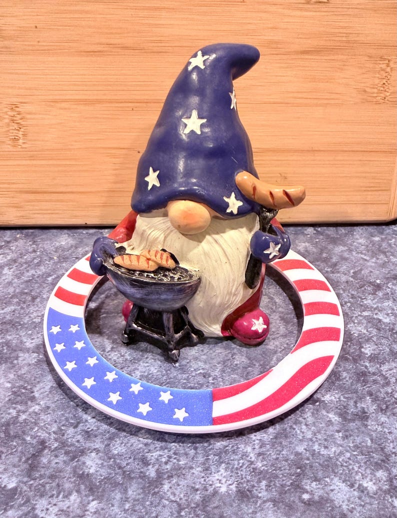 May include: A patriotic gnome figurine with a blue hat adorned with white stars, holding a miniature grill with hot dogs and a sausage. The base is a ring with red and white stripes and blue stars.