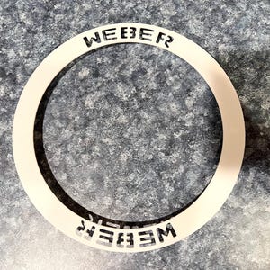 May include: White circular ring with the word "WEBER" cut out in black lettering on the top and bottom of the ring.