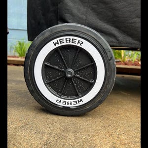 May include: Close-up of a black Weber grill wheel with a white rim. The word "WEBER" is printed in white on the rim. The wheel is on a gray surface.